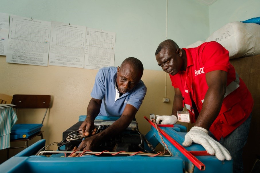 Coca-Cola Lead Cooler Technician, Maxwell Ayisi (right), and Ghana Health Service Refrigeration Technician, Livingstone Modey, repairing a dual gas/ electric vaccine refrigerator at a clinic in Peki Dzake, Ghana. Image by The Coca-Cola Company